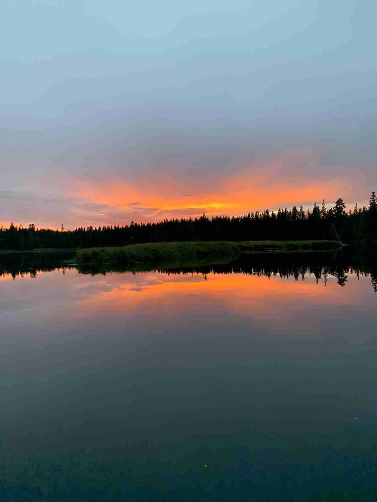 En stilla sjö i Yttermalung speglar en färgrik himmel vid solnedgång, med skogens silhuetter på horisonten.