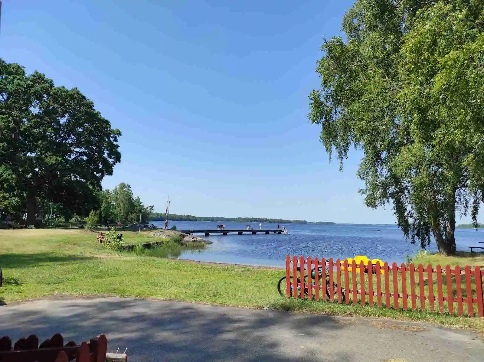 Bild av Hätteboda Vildmarkscamping visar en naturskön sommarvy med en sjö omgiven av gräs och skog. En vattendrag sträcker sig genom landskapet, med en kustlinje som skapar en fridfull atmosfär.