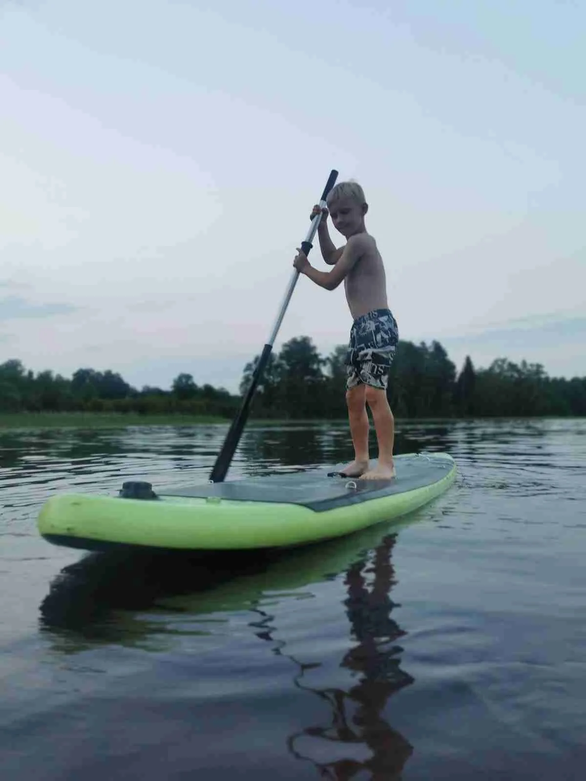 En person paddlar stående på en sup-bräda i en sjö vid Österby Camping, omgiven av grönskande natur och lugnt vatten.