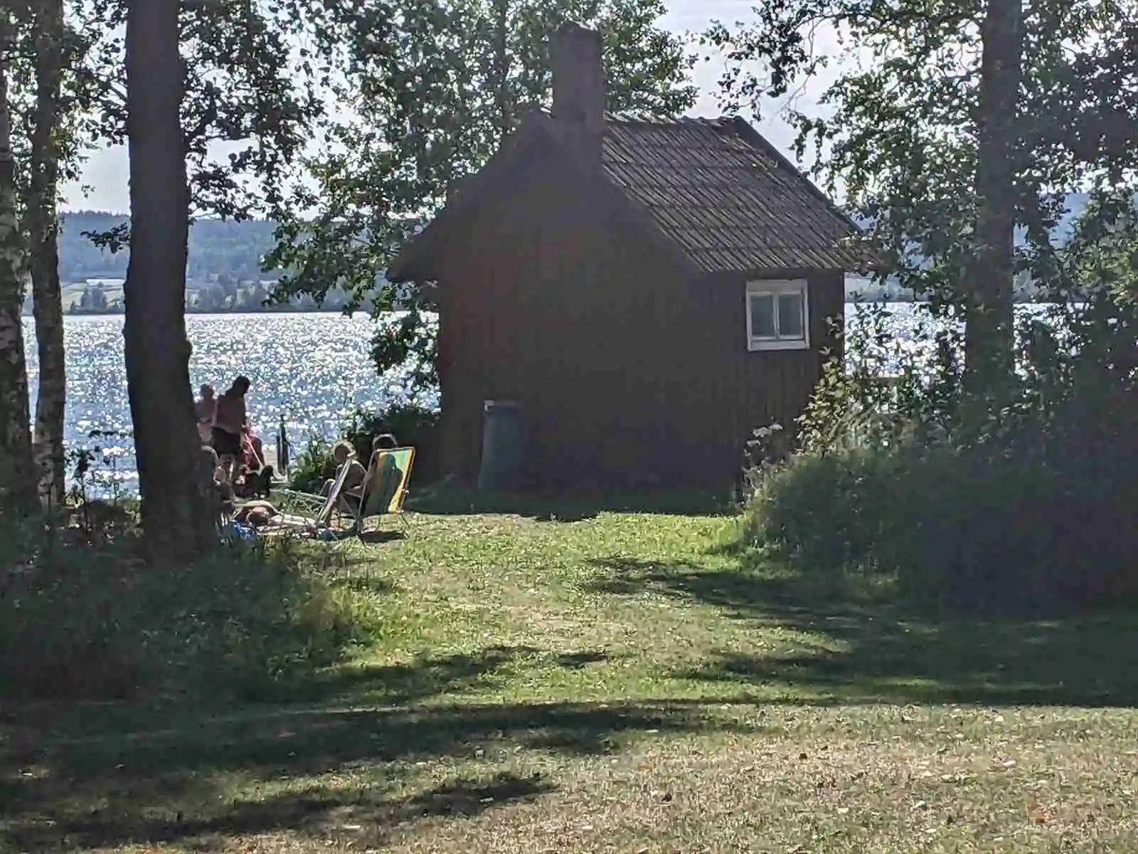 En pittoresk kursgård intill en sjö, omgiven av grönskande gräsmattor och skog. En stuga syns i bakgrunden nära strandkanten.