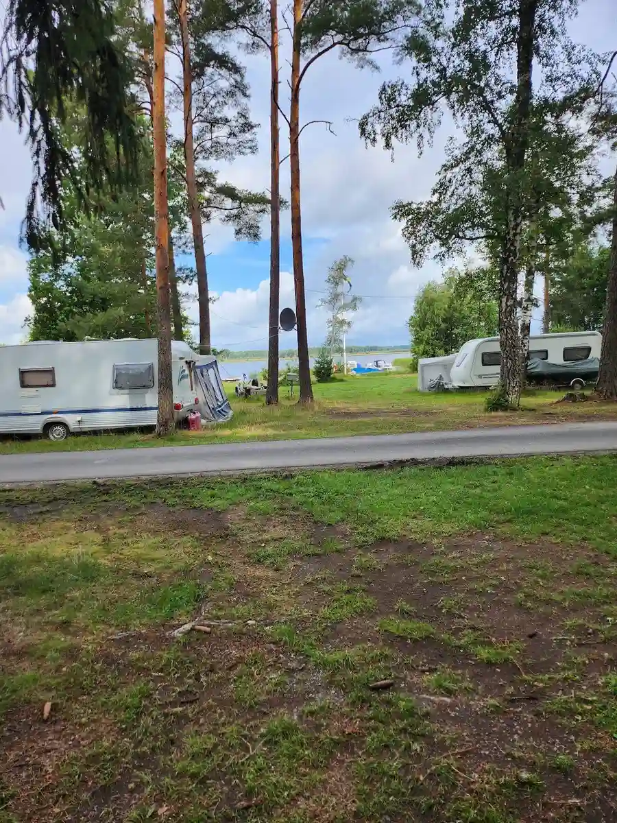 En husvagn står parkerad på E4:ans camping, omgiven av en välklippt gräsmatta och träd. Flera andra campingfordon är synliga i bakgrunden, vilket skapar en inbjudande atmosfär för camping och friluftsliv.