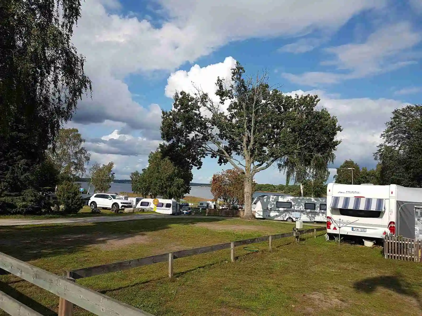 En parkeringsplats vid Ronneby Havscamping med flera olika typer av fordon, inklusive husbilar, familjebilar och lyxbilar, omgivna av en grön parkmiljö.