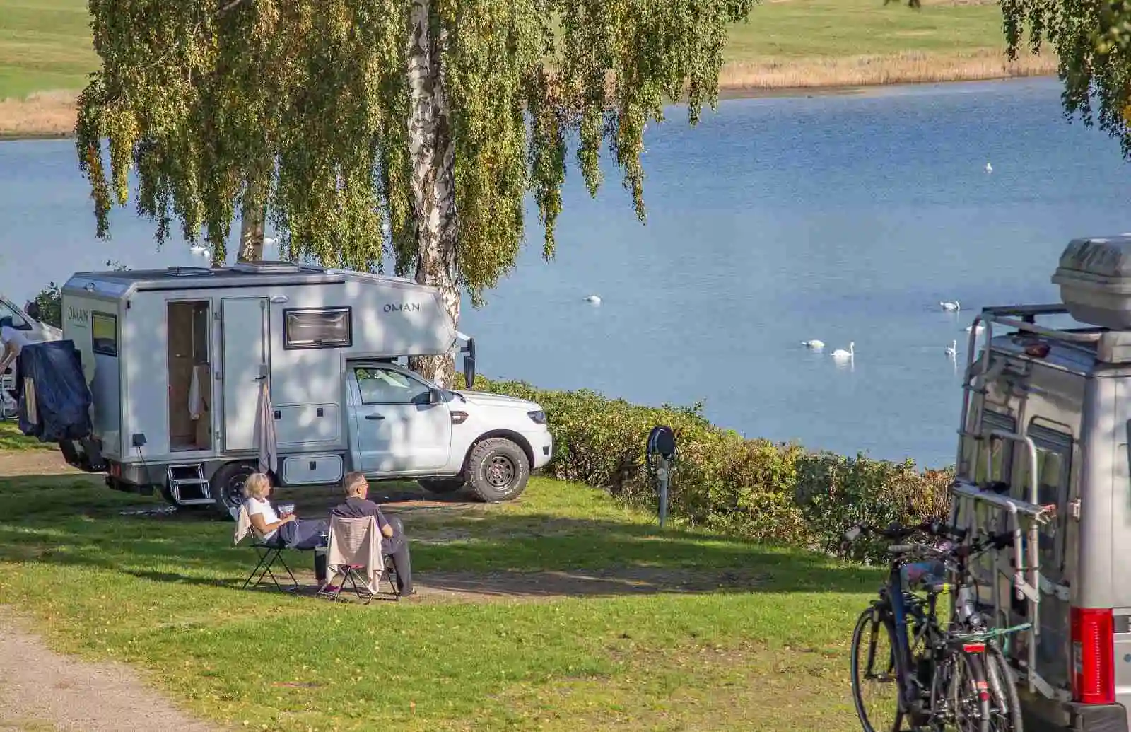En husbil och en husvagn står parkerade på en campingplats med grönska i bakgrunden, nära en sjö eller ett vattendrag. Det är en solig dag, och området ger en inbjudande känsla av friluftsliv och avkoppling.
