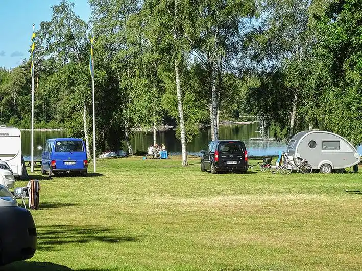 En husvagn står parkerad på en grön gräsyta vid Alsterbro camping, omgiven av flera bilar och ett rymligt parkutrymme.