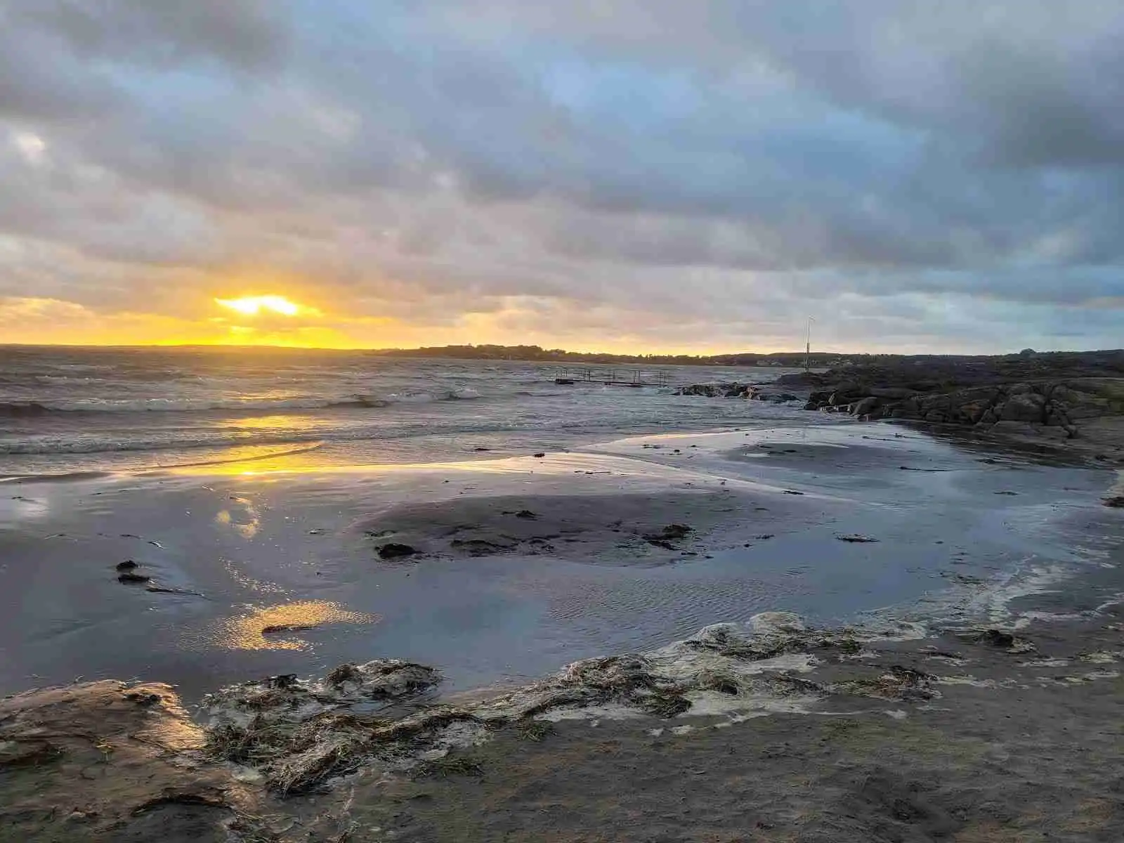 En naturskön kustlinje vid Vallersvik Camping and Hostel med blå himmel, mjuka moln, och en horisont över vattnet vid skymningen. Klippor och en strandlinje syns i förgrunden.