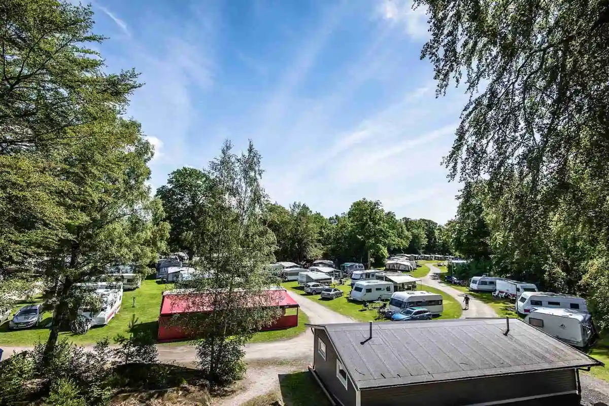 En campingplats med flera parkerade husbilar och bilar på grusunderlag, omgiven av grönska.