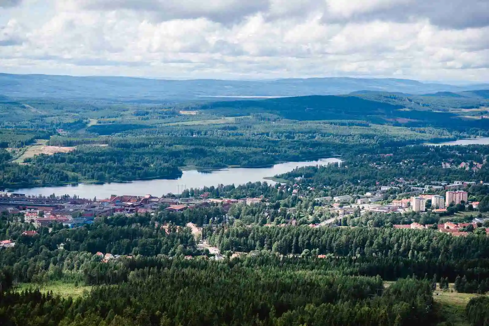 Fågelperspektiv över Alevi camping med böljande kullar, berg i bakgrunden och en sjö i närheten, omgiven av grönska.
