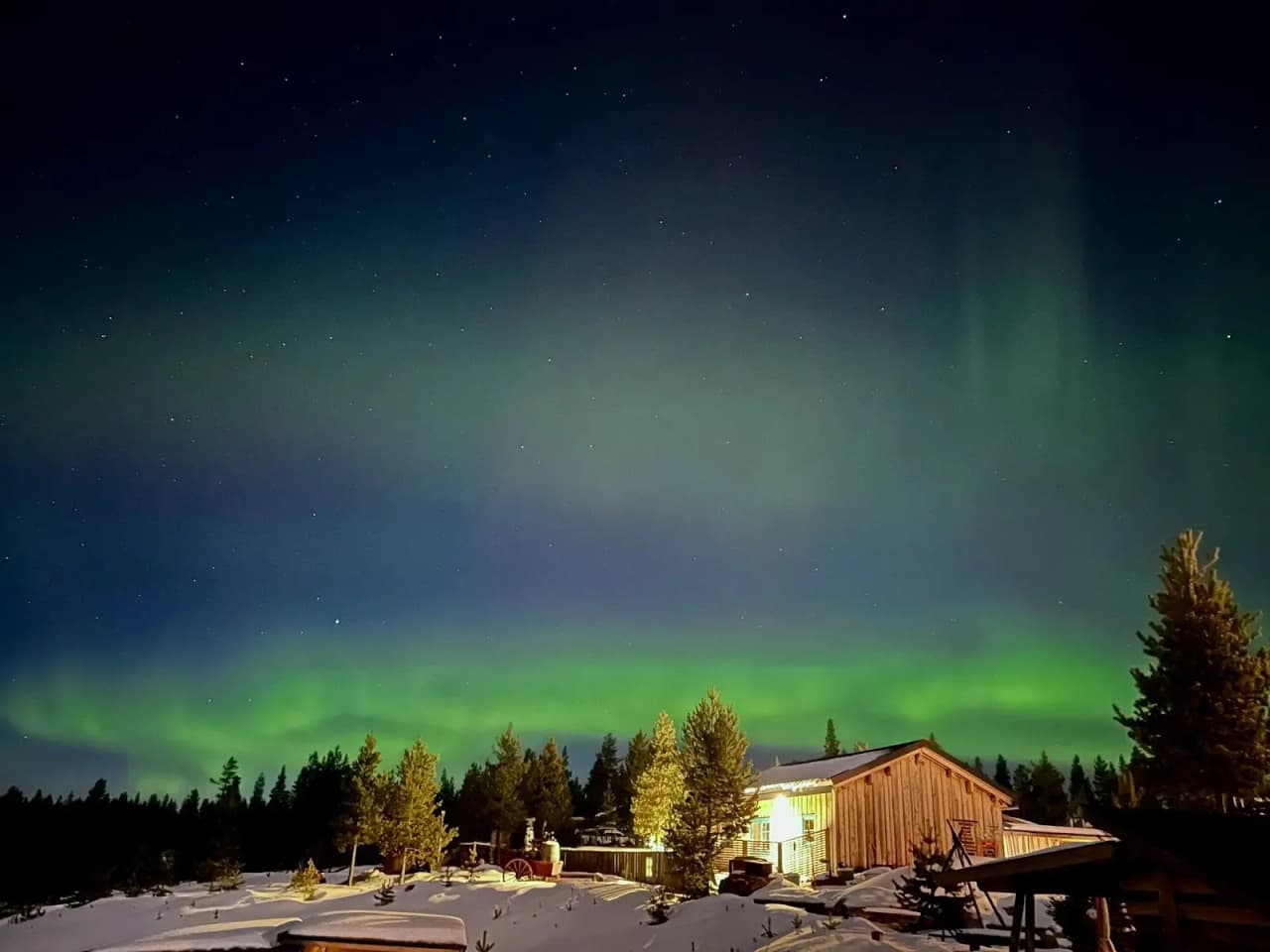 Ett stämningsfullt nattlandskap vid offroadcamp med norrsken som täcker himlen. Ett litet vinterbonat hus står omgivet av snö, med stjärnor som glimmar ovanför i en klar vinternatt.