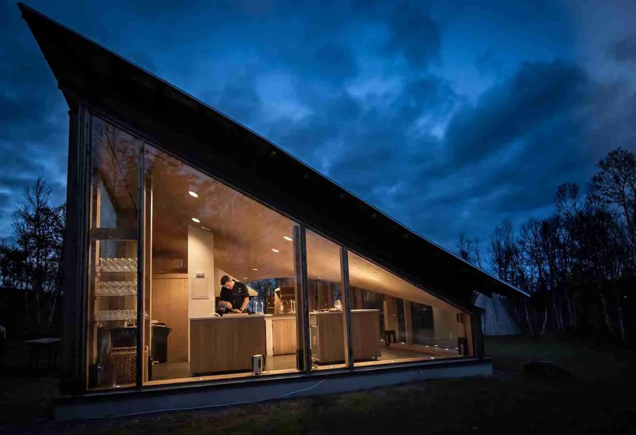 En mysig timmerstuga vid skymning omgiven av grönska, med en lada i bakgrunden och ljuset som faller mjukt på byggnaderna.