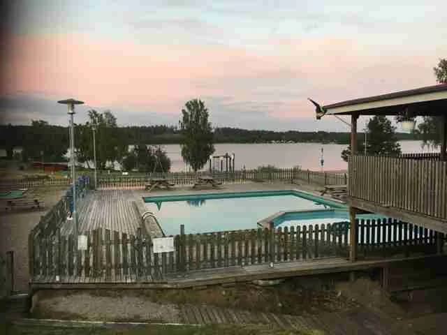En swimmingpool omgiven av solstolar och parasoller, med människor som njuter av avkoppling och fritidsaktiviteter. I bakgrunden syns staket och campinghus, vilket skapar en semesterkänsla på Vegby camping.