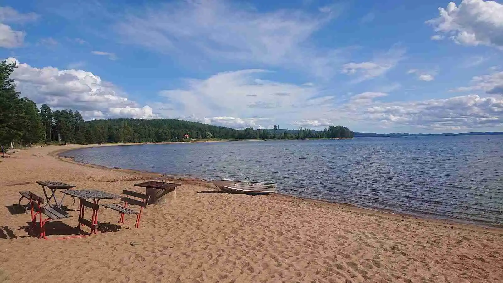 En solig sommardag vid Norra Dellen Familjecamping med en vidsträckt blå sjö, en sandstrand längs kanten och ett naturskönt landskap i bakgrunden.