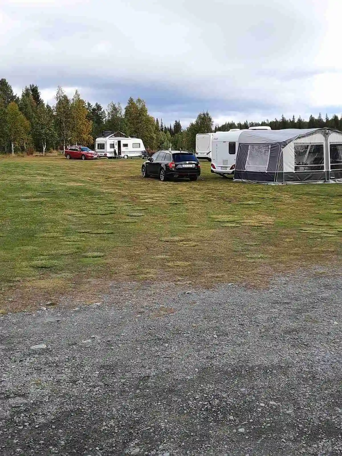 En parkeringsplats vid Emils fiskecamp med olika fordon inklusive husvagnar, familjebilar och tält under en molnig himmel.