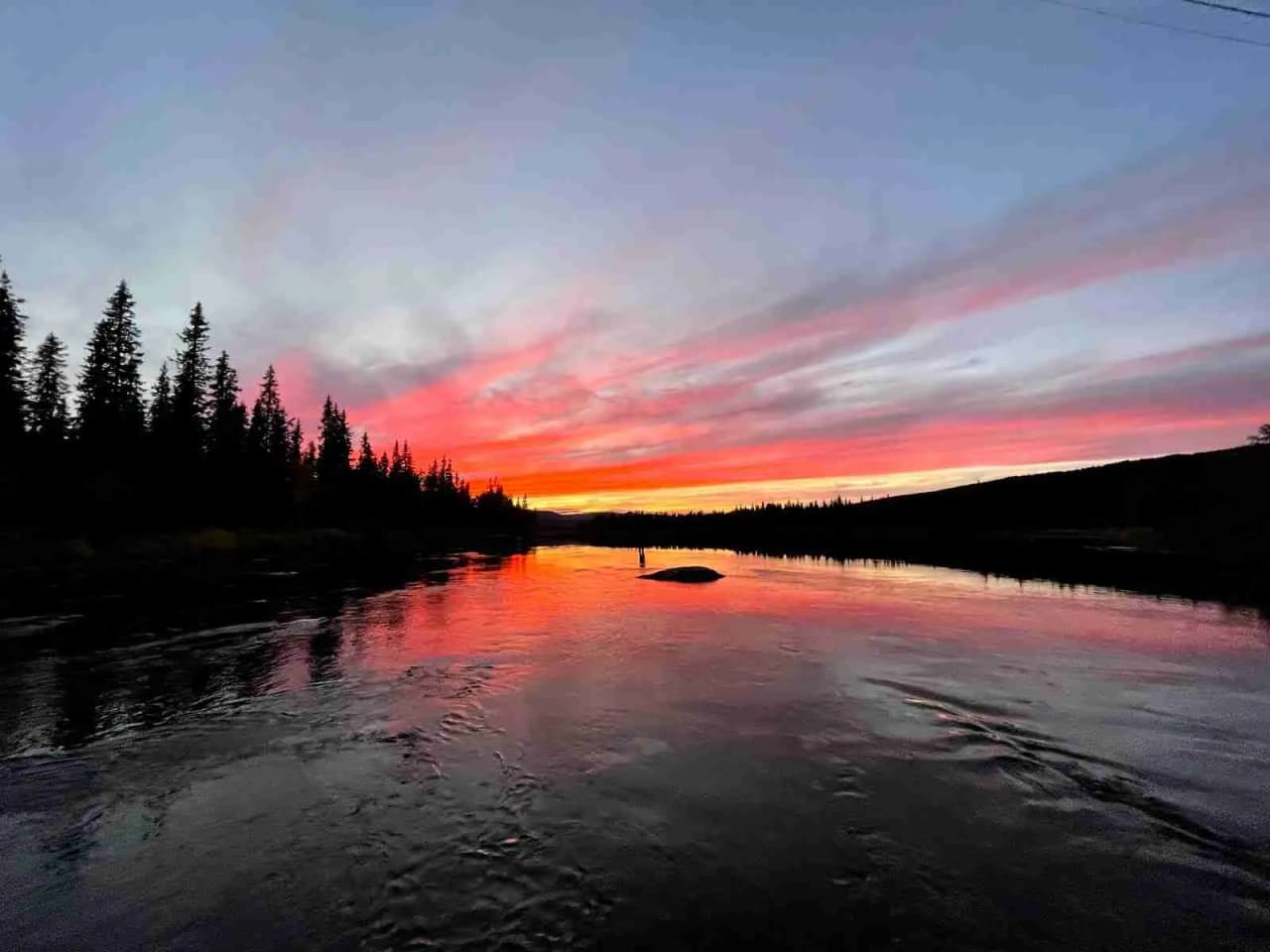 En stillsam solnedgång vid Emils Fiskecamp, med vatten som reflekterar himlens varma färger och moln som svävar över landskapet.