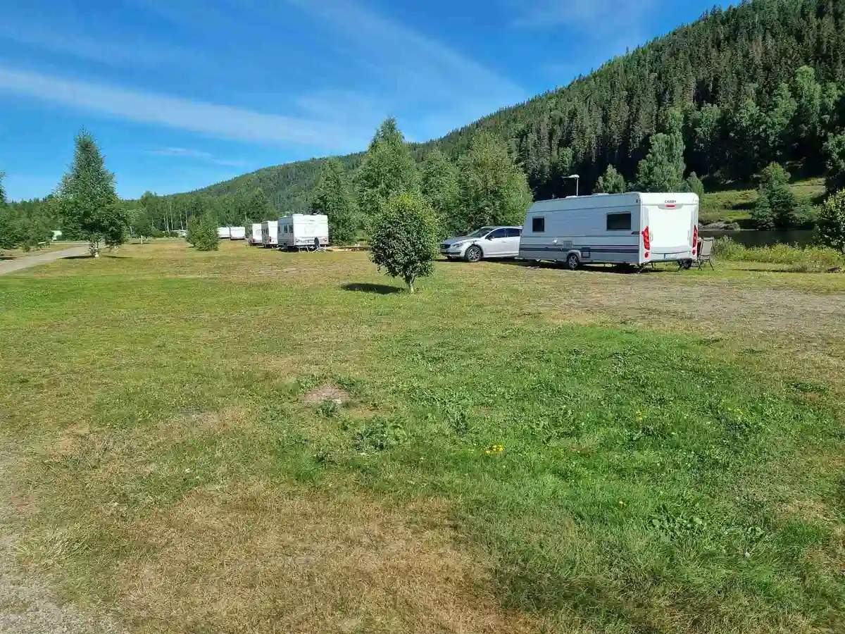 En grön campingplats med husvagnar och husbilar stående på en gräsyta. Omgivningen består av öppna ängar och en lantlig miljö med kullar i bakgrunden.