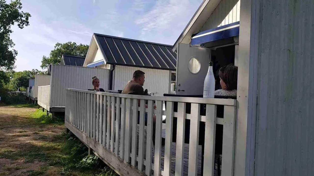 En mysig campingstuga med träpaneler och veranda vid Ronneby Havscamping, omgiven av grönska och trädgårdsbyggnader.