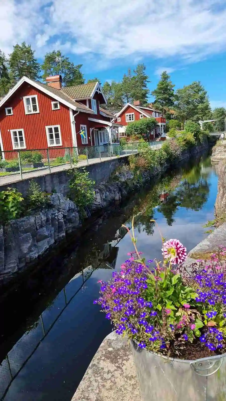 En vattenväg kantad av palmer leder till Elovsbyn Camping & Canoe, med en kanot flytande på spegelblankt vatten. I bakgrunden syns en semesterort omgiven av frodig grönska.