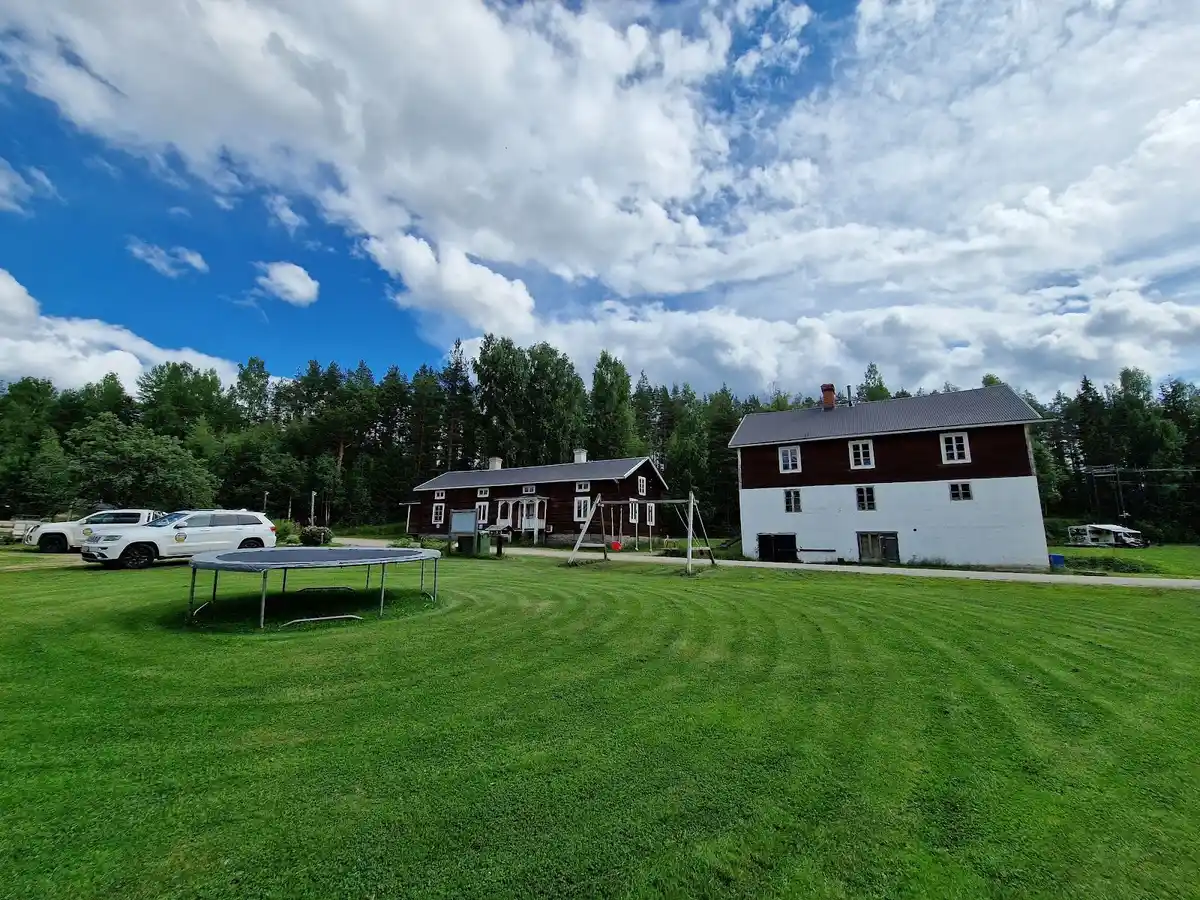 En gräsmatta omgiven av ett öppet landskap med en liten stuga i bakgrunden, omgiven av träd och växter, skapar en naturskön och rofylld miljö vid Camp Ängra i Wildlife Sweden.