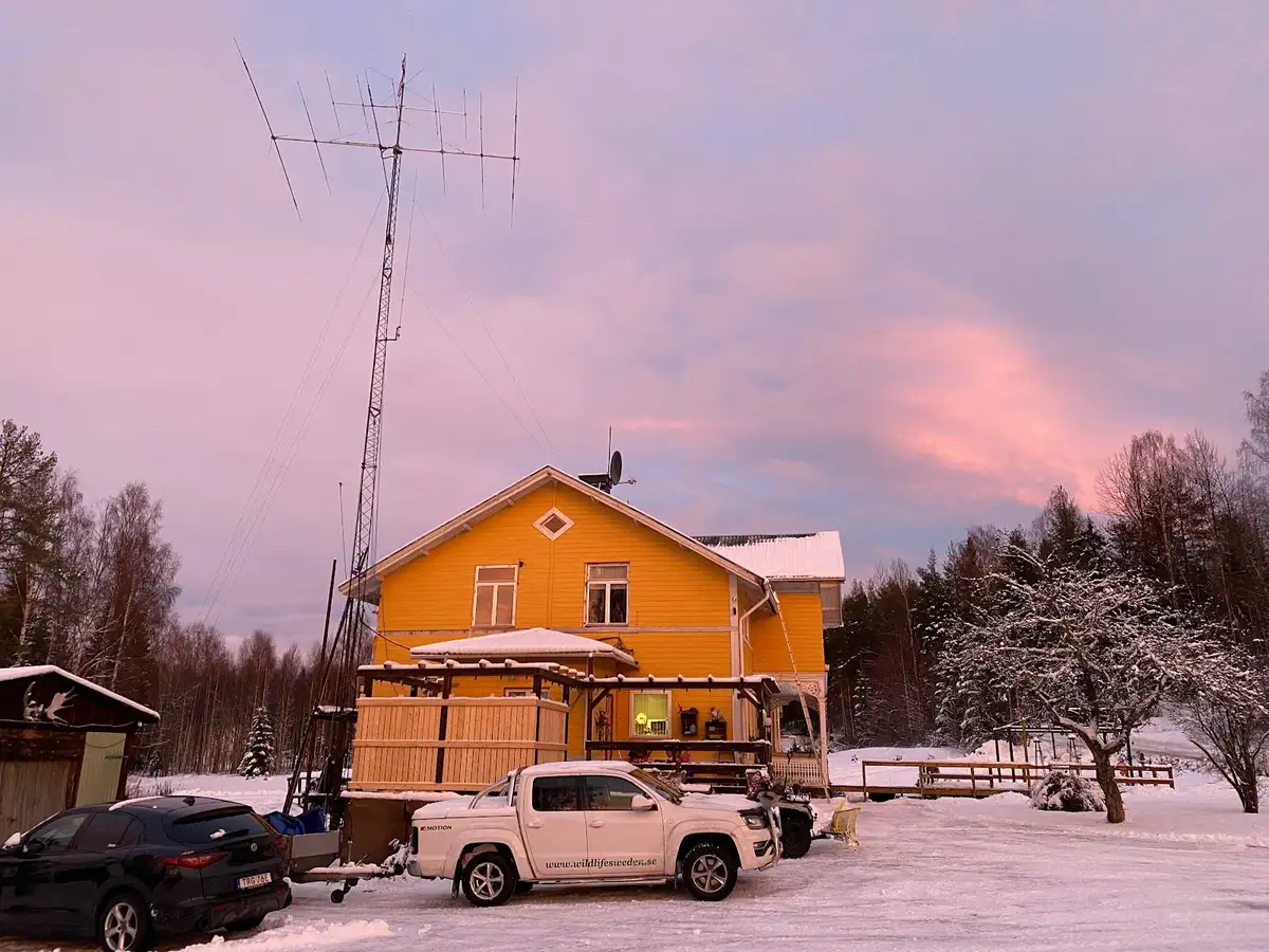 En liten stuga täckt med snö står mitt i ett vinterlandskap vid Wildlife Sweden Camp Ängra, med en sportbil parkad framför och överliggande kraftledningar i bakgrunden.