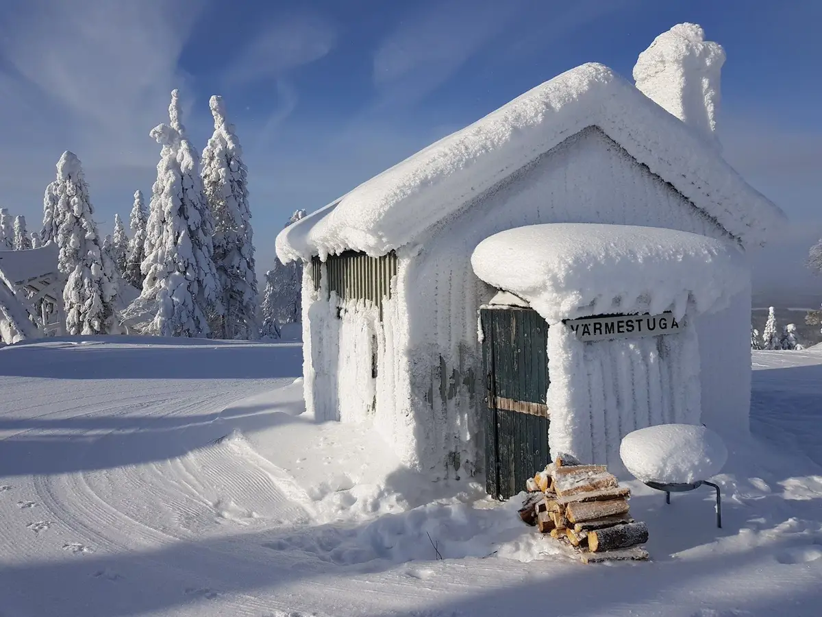 En stuga med snötäckt tak mitt i ett vintrigt landskap vid Storklinta Skidcenter AB. Snön täcker marken och det ser kallt och frostigt ut, idealiskt för vintersport och skidåkning.