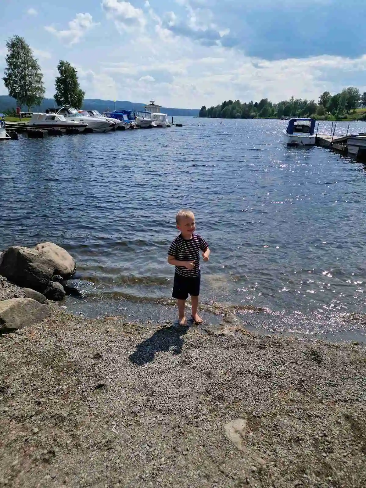 En motorbåt ligger förtöjd vid en sjö med klarblått vatten, omgiven av moln och gröna kustlinjer, vid Hoverbergs camping.