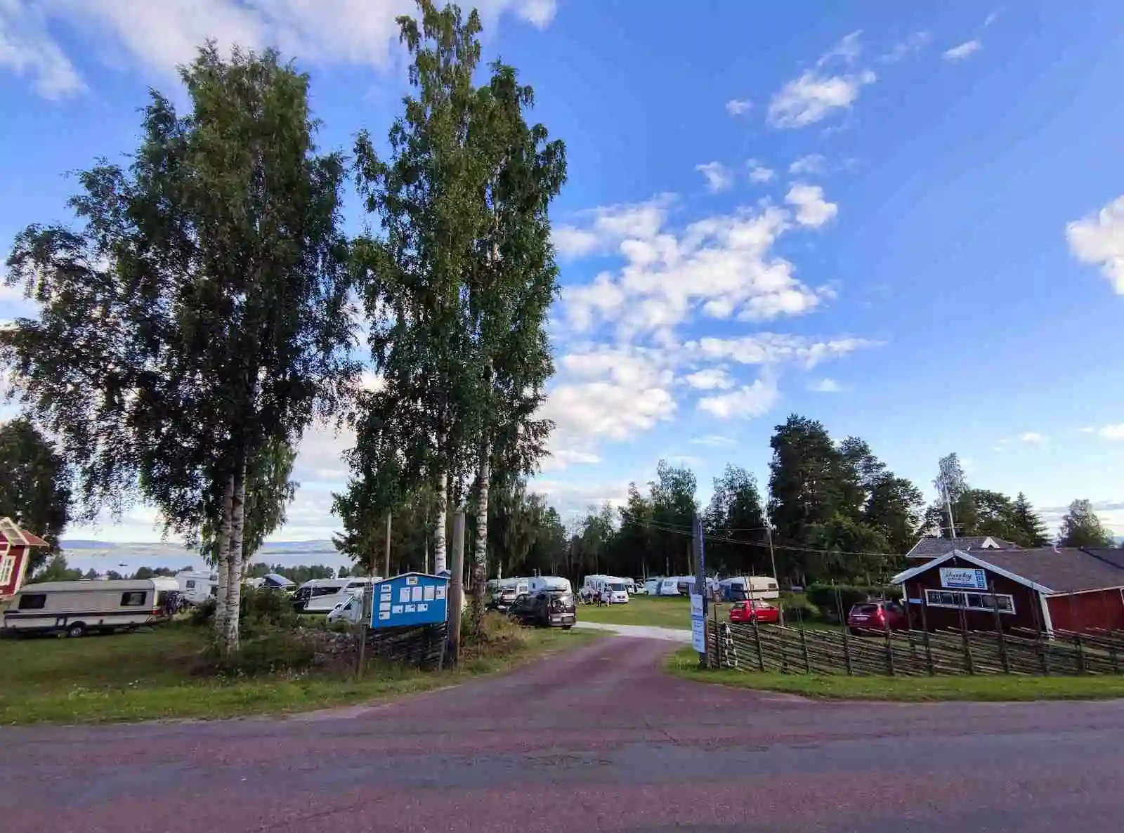 En parkeringsplats vid Vikarbygården med några bilar och husvagnar, omgiven av grönskande gräsmattor och molnig himmel.