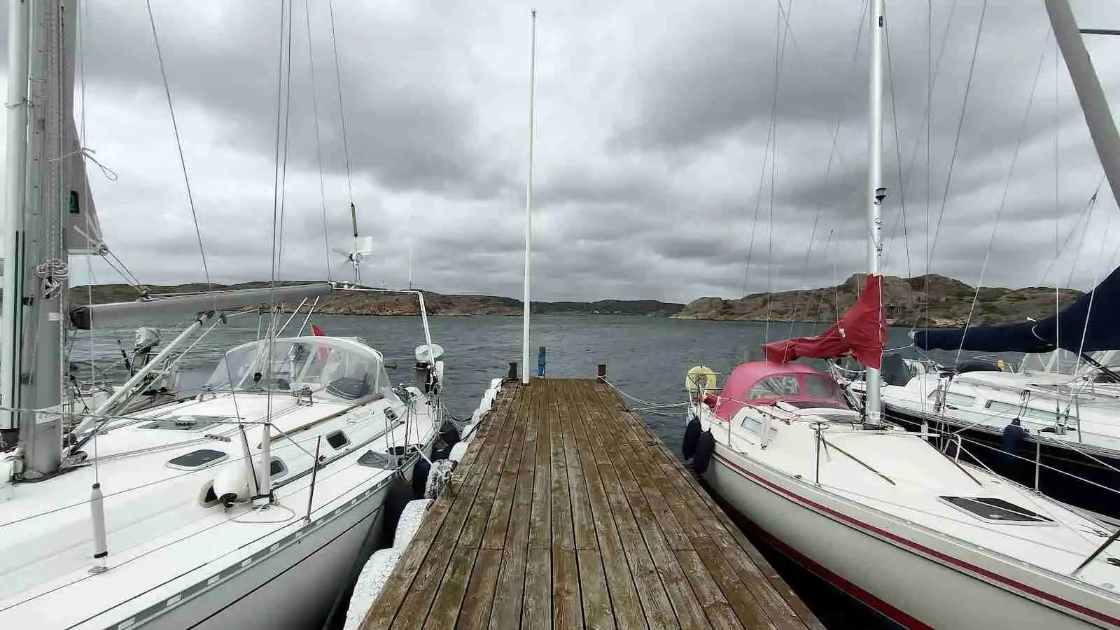 En marinavy med flera segelbåtar och vattenfarkoster förtöjda vid en brygga i hamnen vid Mossholmen. Mastarna reser sig mot en klar himmel, och det omgivande havet är stilla.