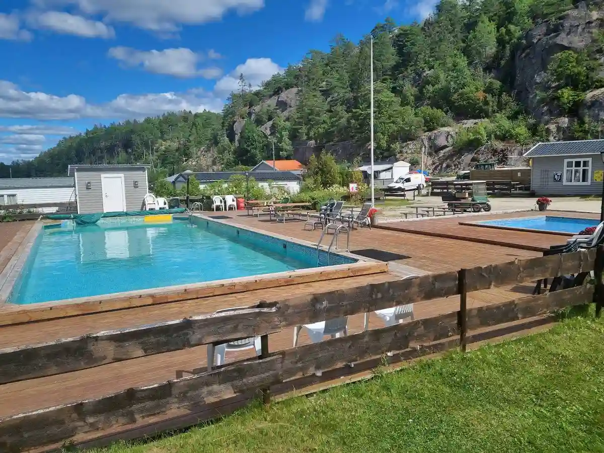 En solig dag vid ett semesterresort med en utomhuspool omgiven av solstolar och utemöbler, beläget i ett naturskönt område.