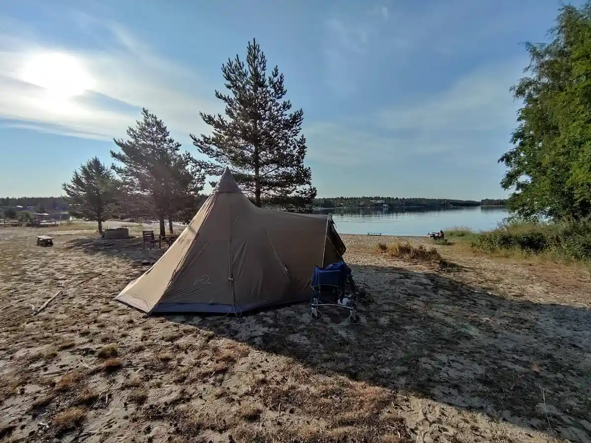 En tältplats vid Ava havsbad och camping med flera tält uppsatta under skuggande presenningar. Utrustning för camping och vandring syns i området.