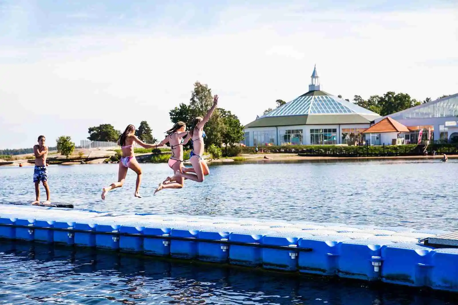 En somrig dag på Västervik Resort med människor som badar och njuter av solen vid vattnet. Besökare roar sig med olika aktiviteter, klädda i badkläder och omgivna av semesterstämning.