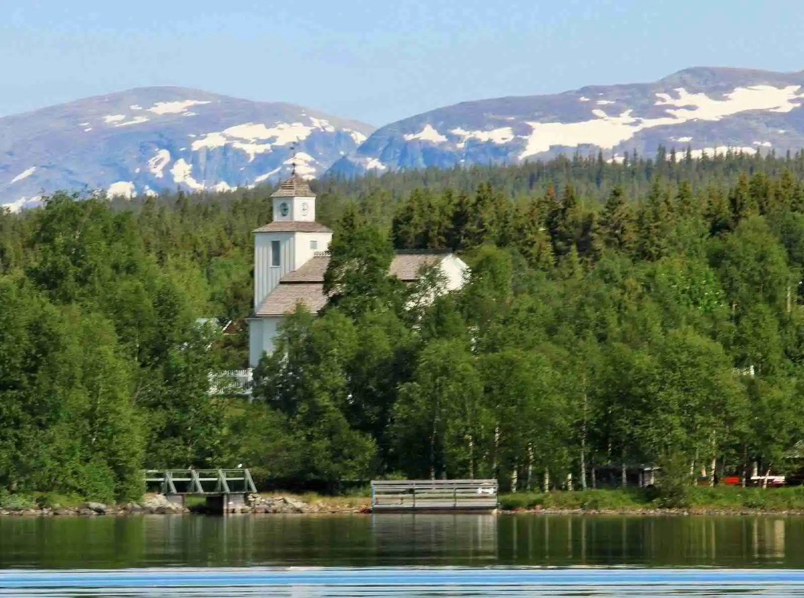 Bild av Storsjö camping och ställplats som visar en naturskön vy med en sjö i förgrunden omgiven av tät barrskog. I bakgrunden syns ett torn likt en kyrktornsspira, vilket ger en känsla av närhet till naturen och stillheten vid en sjö.