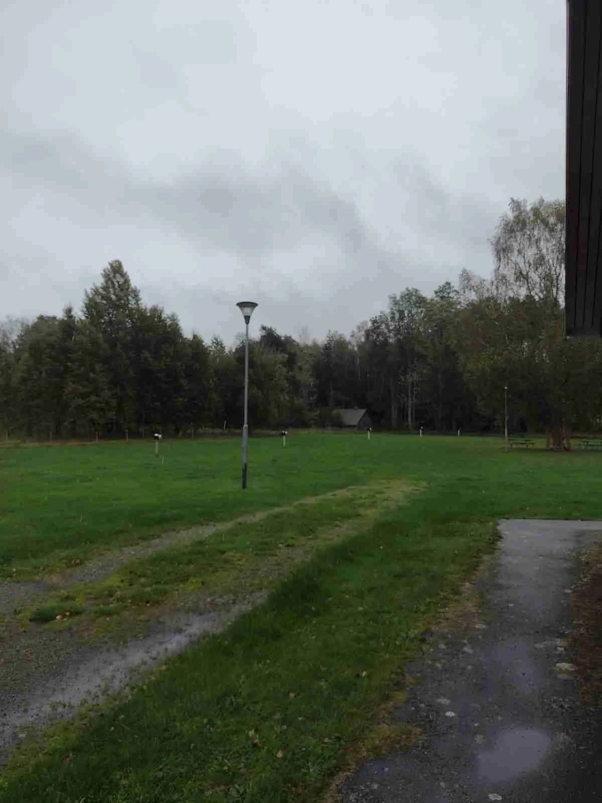 En vidsträckt gräsyta med spridda gräspoler och en flaggstång inbäddad i en naturskön miljö vid Emmaboda camping.