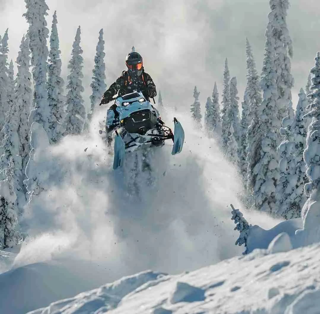 En grupp människor kör snöskoter över ett snötäckt vinterlandskap vid sportcamping Åkersjön, omgivna av snöiga kullar och träd under en klar vinterdag.