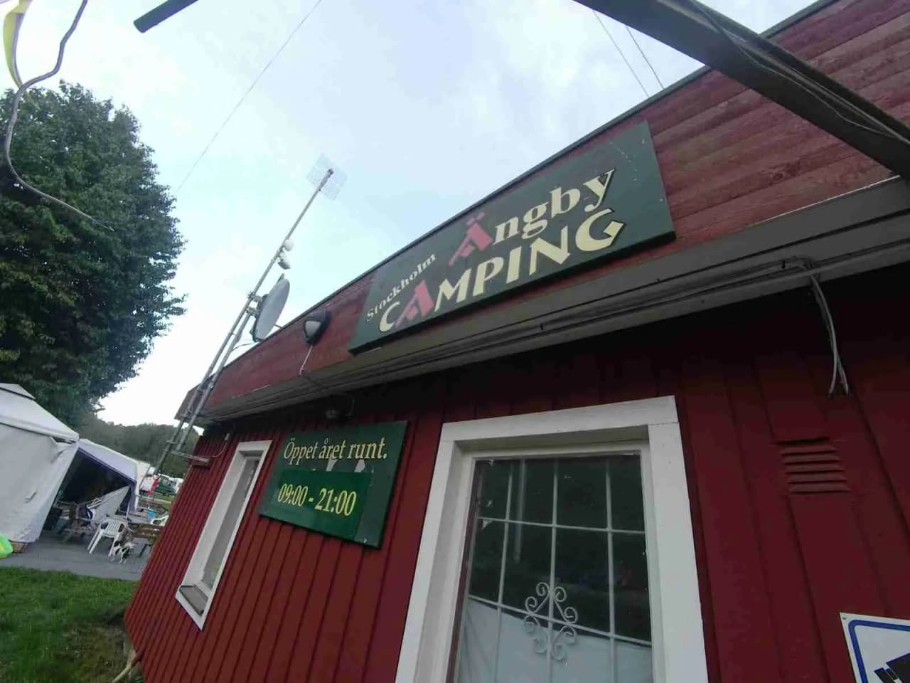 En grupp tält uppställda på ett gräsområde vid Ängby Camping, omgivna av träd och en klar himmel.