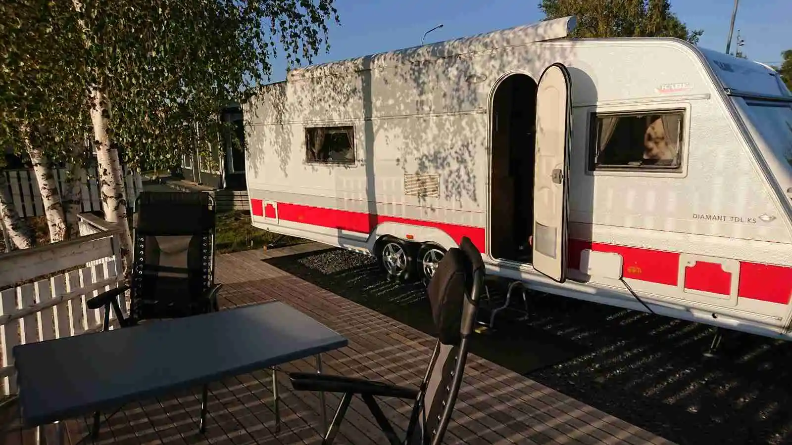 En campingplats med flera husvagnar och husbilar parkerade på gräsytan. I bakgrunden syns några träd och en klar himmel.