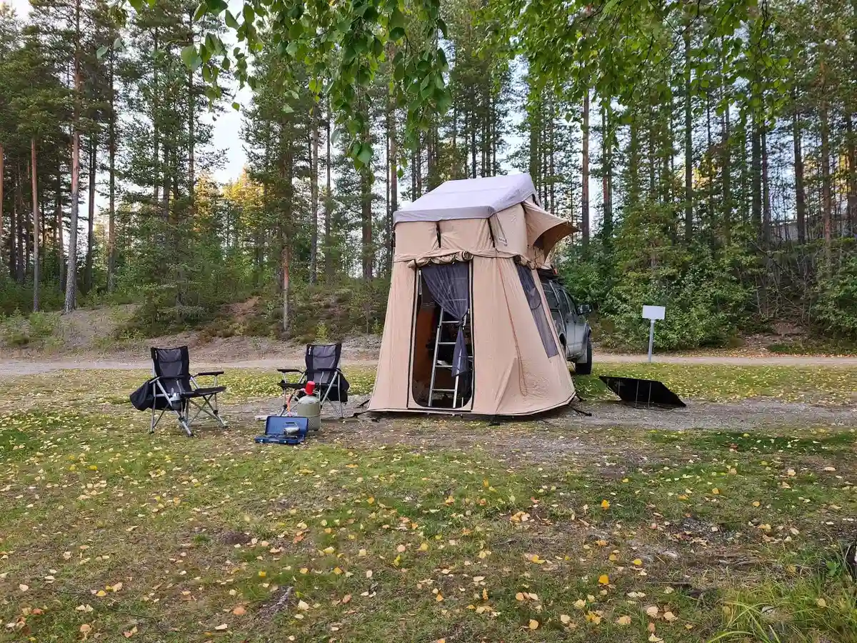 En bild som visar en campingplats vid Lapplandsportens camping med tält och husvagn under ett skyddande presenningstak. I bakgrunden syns naturen som omger platsen, vilket ger en känsla av friluftsliv och rekreation.