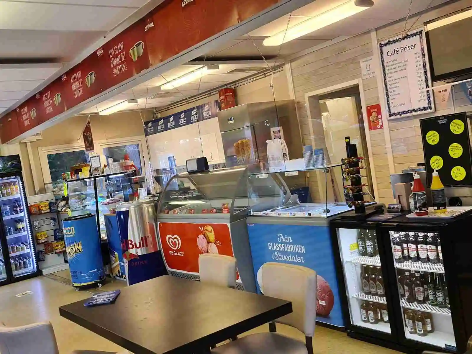 Interiör av Gävle Camping Engesberg med modern design och mjuk belysning. Taklampor och ljusa golv präglar rummet, där ett litet caféområde med en TV och logotyp är i fokus. Restaurangmiljön ger en inbjudande och avslappnad atmosfär.