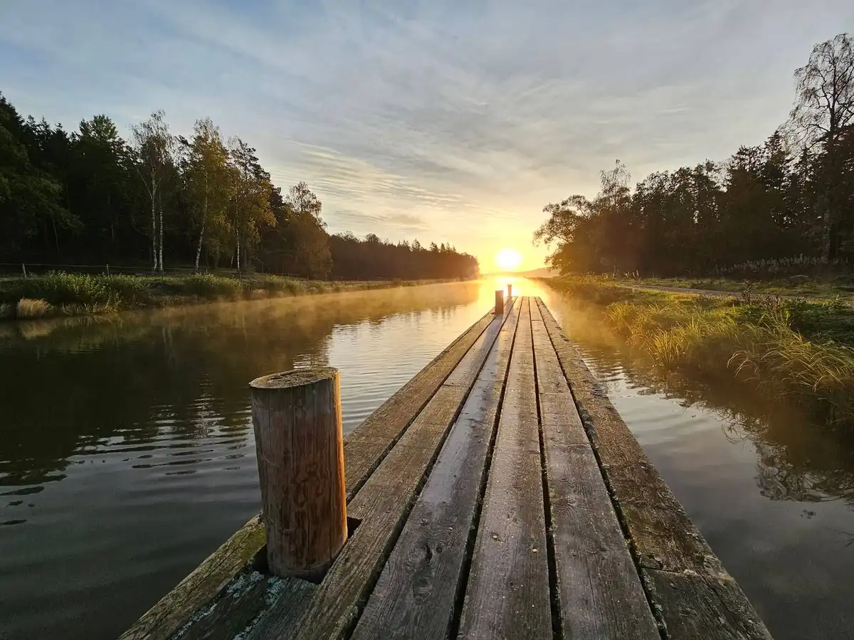 En fridfull camping vid en vattenstig, kantad av träd och grönska, med en träbrygga i förgrunden. Morgonljuset speglas i vattnet, omgärdat av moln på himlen.