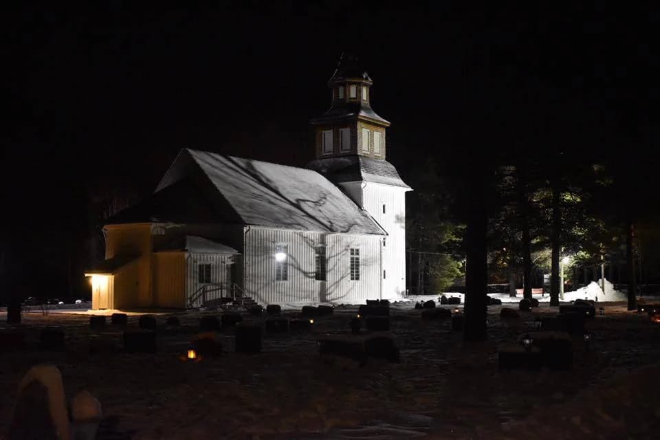 En mörk nattscen visar en kyrka med ett högt klocktorn, omgiven av ett klosterliknande byggnadskomplex. Allt är stilla och tyst under en stjärnklar himmel i kvällsmörkret.