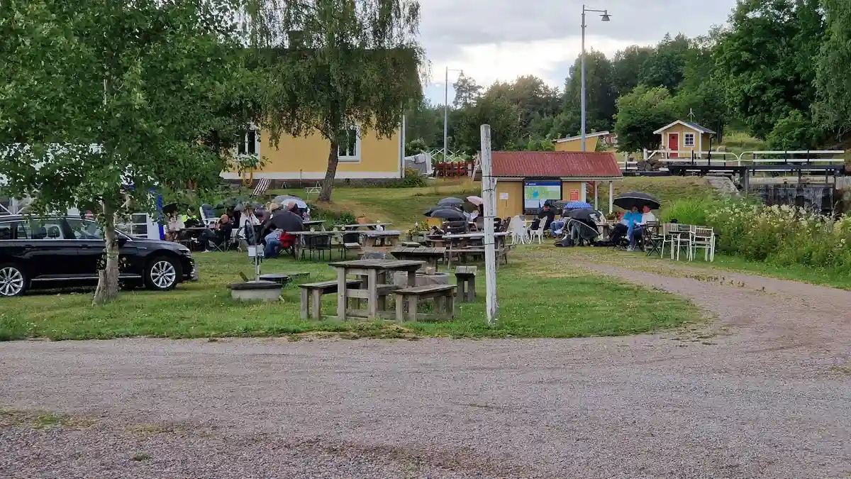 En grönskande campingplats med picknickbord och bänkar, omgiven av en välklippt gräsmatta och parkeringsplats i närheten.