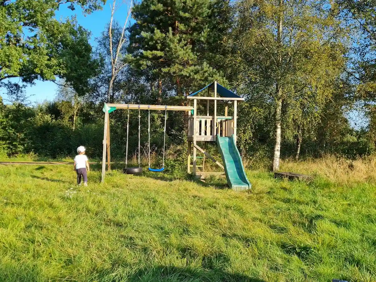 En grönskande äng med en enkel lekplats innehållande en rutschkana, omgiven av öppna gräsmarker på Hallsjö Vildmarkscamping.