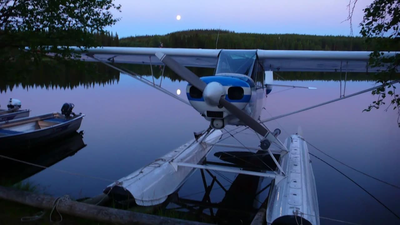 Ett sjöflygplan står stilla vid en brygga vid Rörvattnets Fiskecamp, omgiven av ett naturskönt landskap med sjö och skog i bakgrunden.