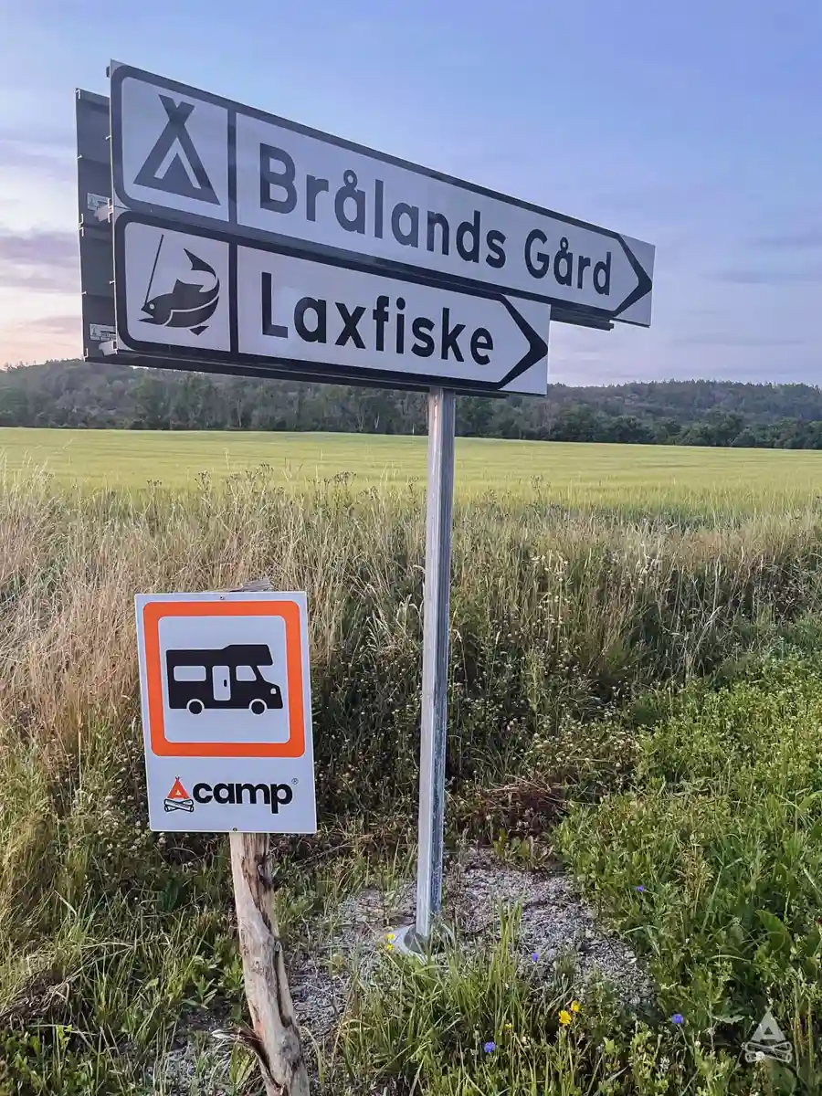 En träskylt vid en grusväg visar vägen till Brålands Gård Nature Camp, omgiven av vidsträckta ängar och natursköna vyer.