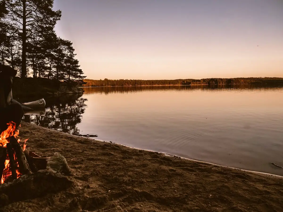 En fridfull skymningsscen vid Viking Republic Nature Camp med vatten i förgrunden, skogsklädd strandlinje och vildmark i bakgrunden.
