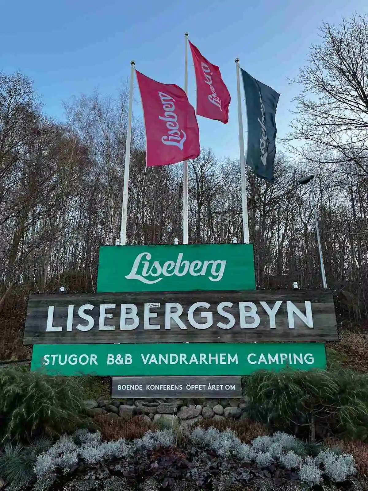 Reklamskylt för Lisebergsbyn i en parkmiljö, omgiven av grönska och träd.