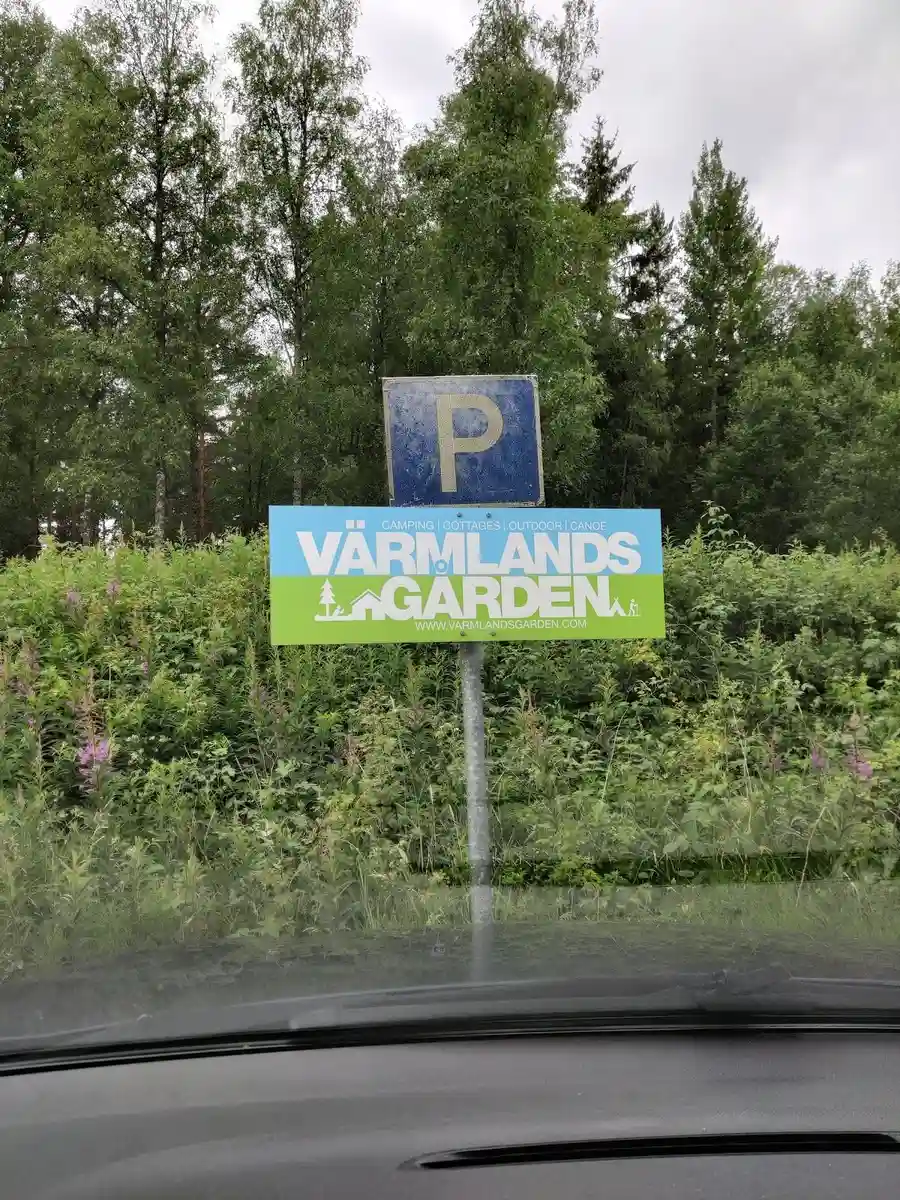 En skylt nära en grusväg omgiven av buskar, som visar "Värmlandsgården" i bakgrunden.