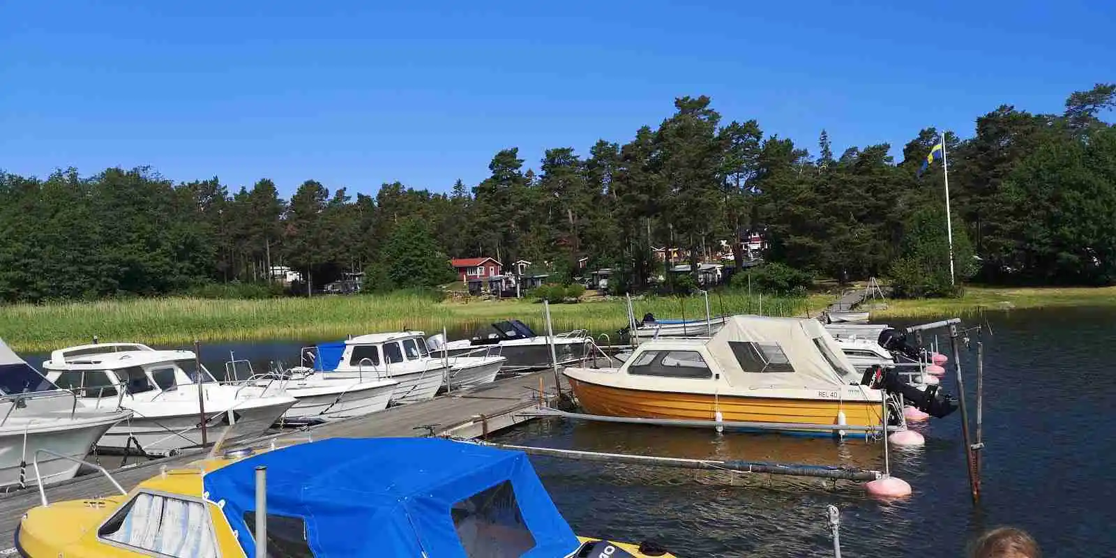 En motorbåt förtöjd vid en brygga i en småbåtshamn med flera andra båtar, omgiven av lugna vatten och grönskande natur vid Norrtälje Camping.