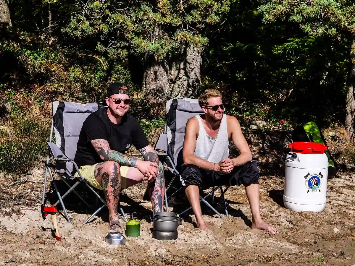 En naturskön campingplats med sommarmiljö, där besökare njuter av fritiden. Bilden visar personer i shorts och badbyxor som kopplar av på vikbara stolar, omgivna av resväskor och väskor, vilket förstärker semesterkänslan i denna utomhusmiljö.