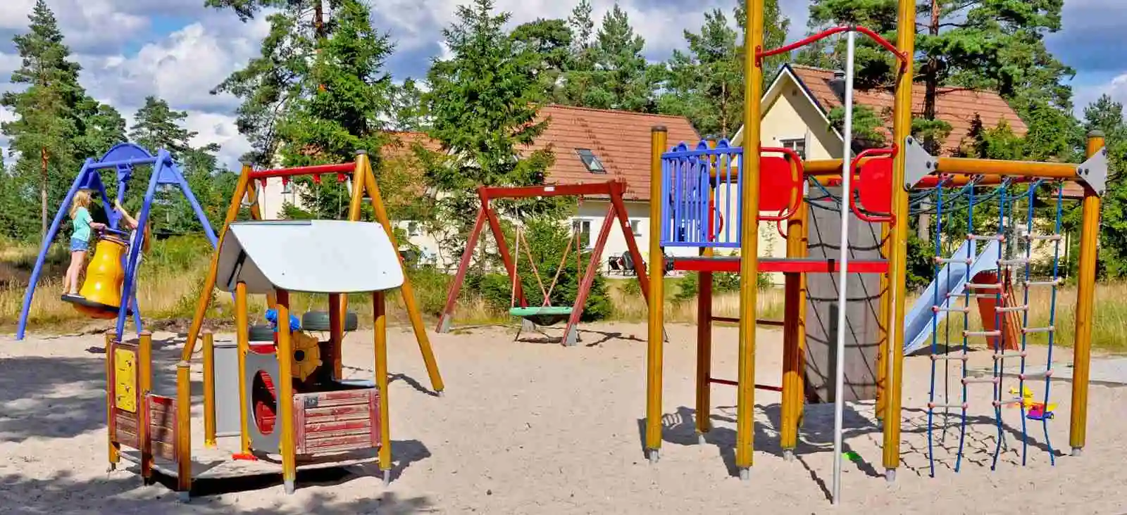 En lekplats utomhus med en stor rutschkana och andra lekanordningar. Omgiven av gröna ytor och parkmiljö, är det en plats för rekreation och lek för barn och familjer.