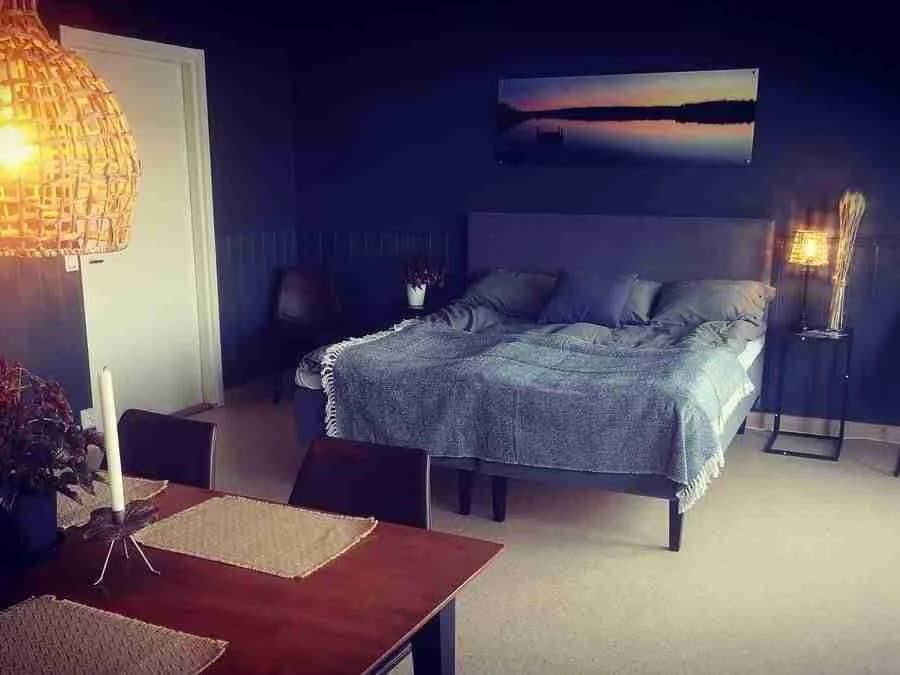 Bild på ett stilrent inrett sovrum med ljusa textilier och trädetaljer. Rummet har en dubbelsäng, träparkettgolv och mjuk belysning, vilket ger en hemtrevlig och mysig atmosfär.