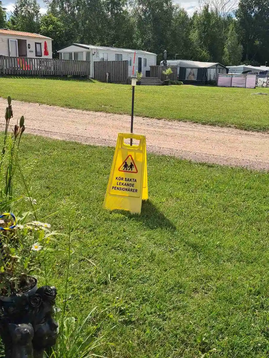 Bild av Ivö Camping med en stor gräsyta i förgrunden. I bakgrunden ses en skylt som markerar området. Det finns också några konformade objekt, sannolikt trafik- eller säkerhetskonor, och omgivningen ger en känsla av en lugn parkmiljö.